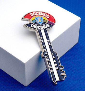 Key-shaped lapel pin featuring the Latin phrases Docendo and Discimus with a blue, red, and purple crest design, placed on a white block over a blue background.