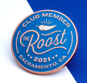 Circular blue and copper lapel pin for The Roost Club Member 2021 in Sacramento, CA, featuring a feather illustration.