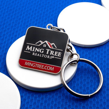 Ming Tree Realtors custom metal keychain with a square black and red enamel logo design, displayed on round white stands over a blue textured background.
