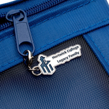 Custom silver zipper pull with Hartwick College Legacy Family logo attached to a blue fabric bag.