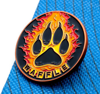 Circular enamel lapel pin with a black paw print surrounded by flames and the name Waffle in yellow lettering.