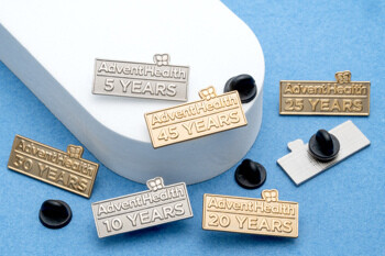 A collection of gold and silver rectangular lapel pins for AdventHealth marking 5, 10, 20, 25, 30, and 45 years of service, displayed on a blue and white surfac