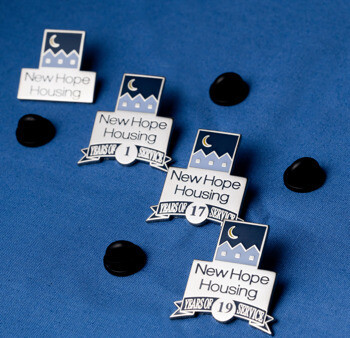 Custom New Hope Housing years of service lapel pins with housing logo and black rubber clutches on blue fabric.