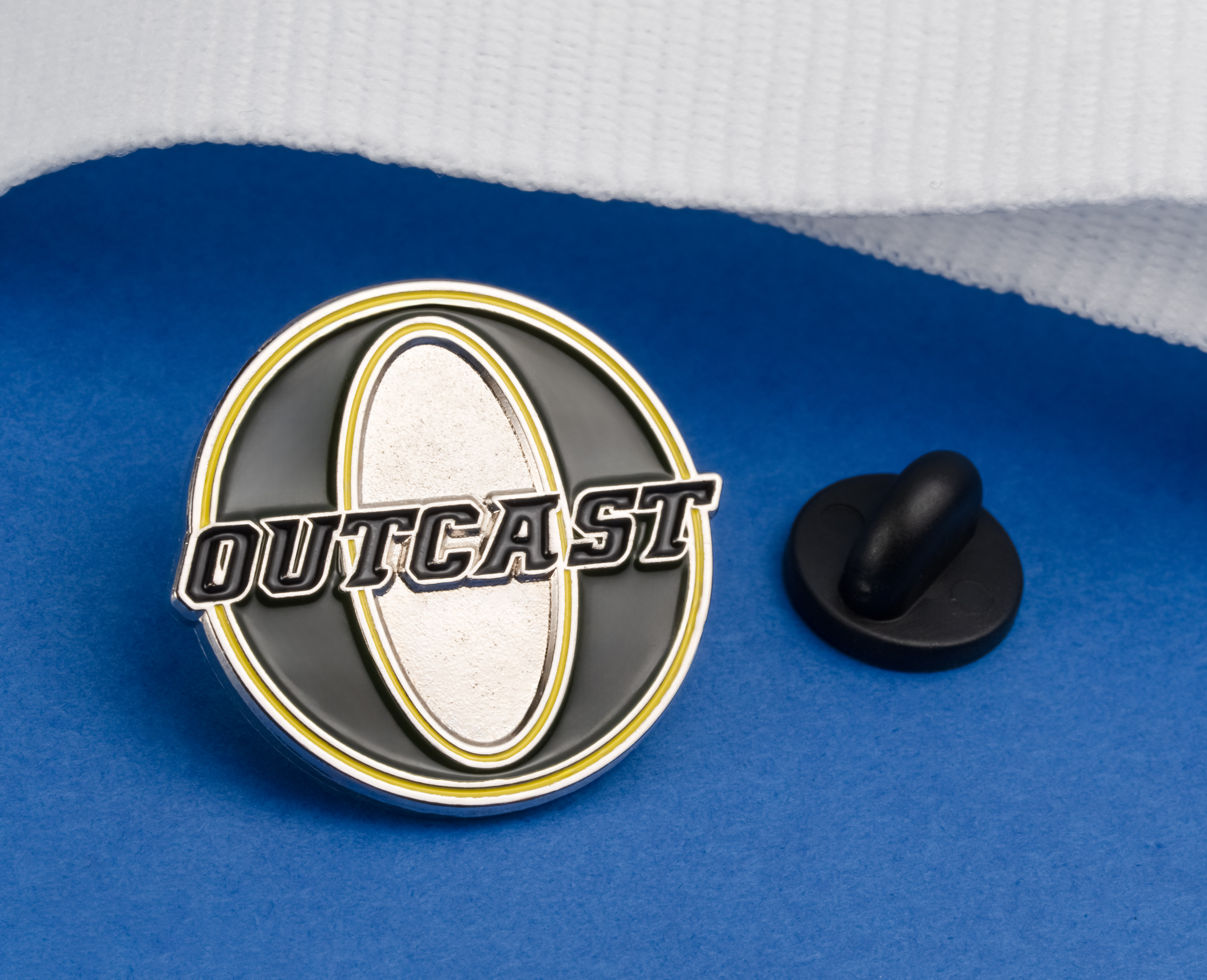 Circular black and silver lapel pin featuring the word Outcast over a silver oval with yellow accents and a black rubber clutch.